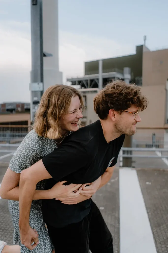 Paarshooting im Parkhaus Münster Junges Paar lacht, während die Frau den Mann von hinten umarmt.