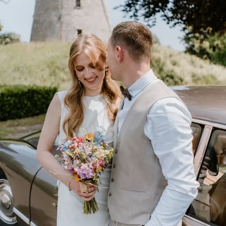Brautpaarshooting mit Oldtimer bei Mühle Ennigerloh bei Münster Braut mit Blumenstrauß und Bräutigam, vor einem Auto und einer Mühle im Freien.